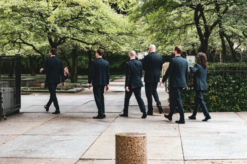 Groomsmen in matching dark suits walking away through a leafy plaza, seen from behind